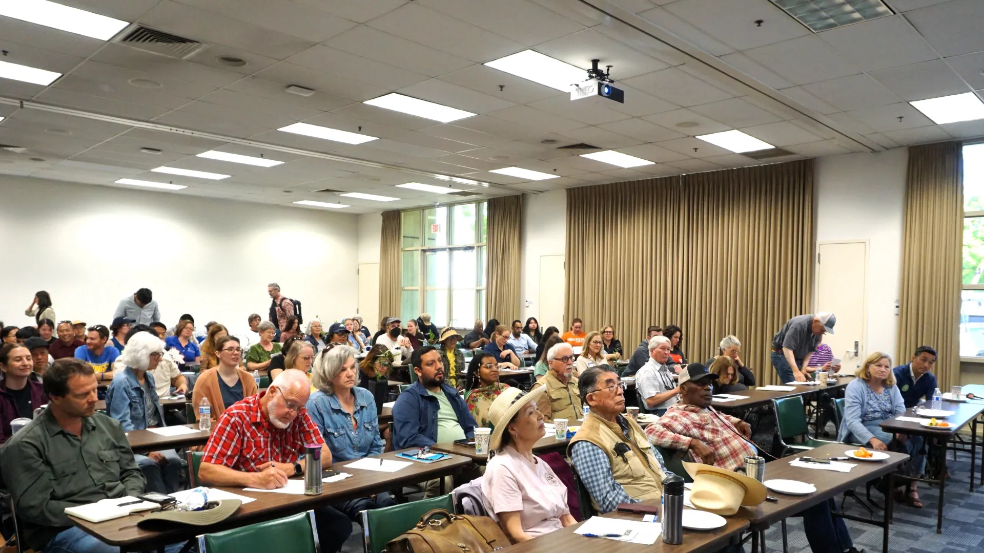 Rows of people sit at long tables in a large seminar room, listening to a speaker during Tea Day