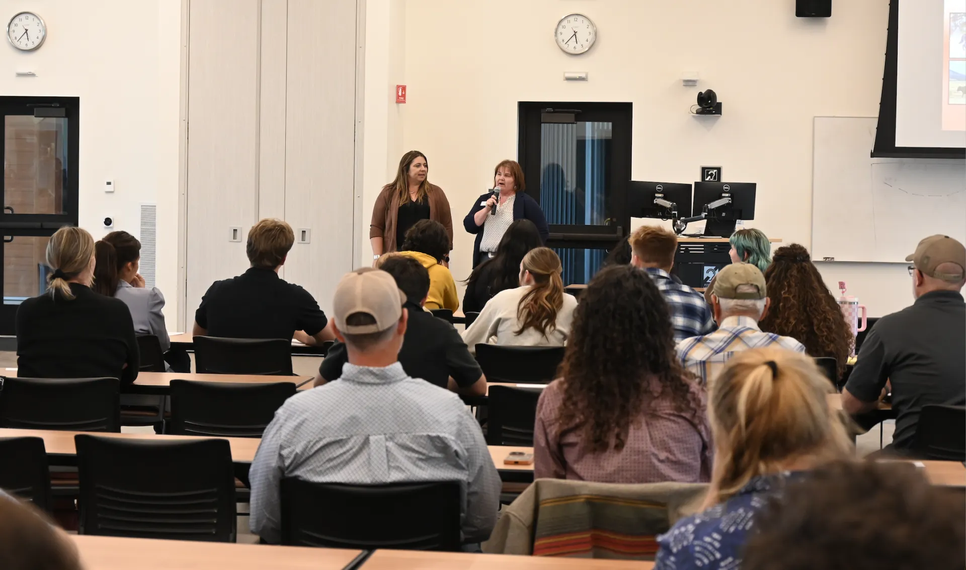 Tracy, left, stands beside Kasey, who is speaking into a mic as they face the audience