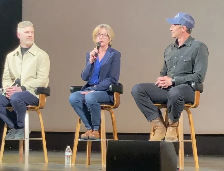 A woman seated on a stool onstage speaks into a mic as men on either side of her watch her speak
