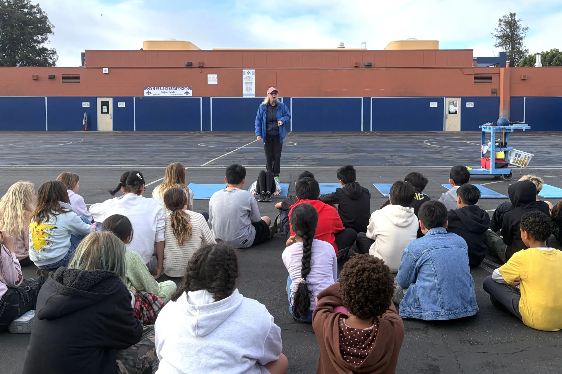 A physical education teacher addresses a class of fifth graders sitting on school yard blacktop