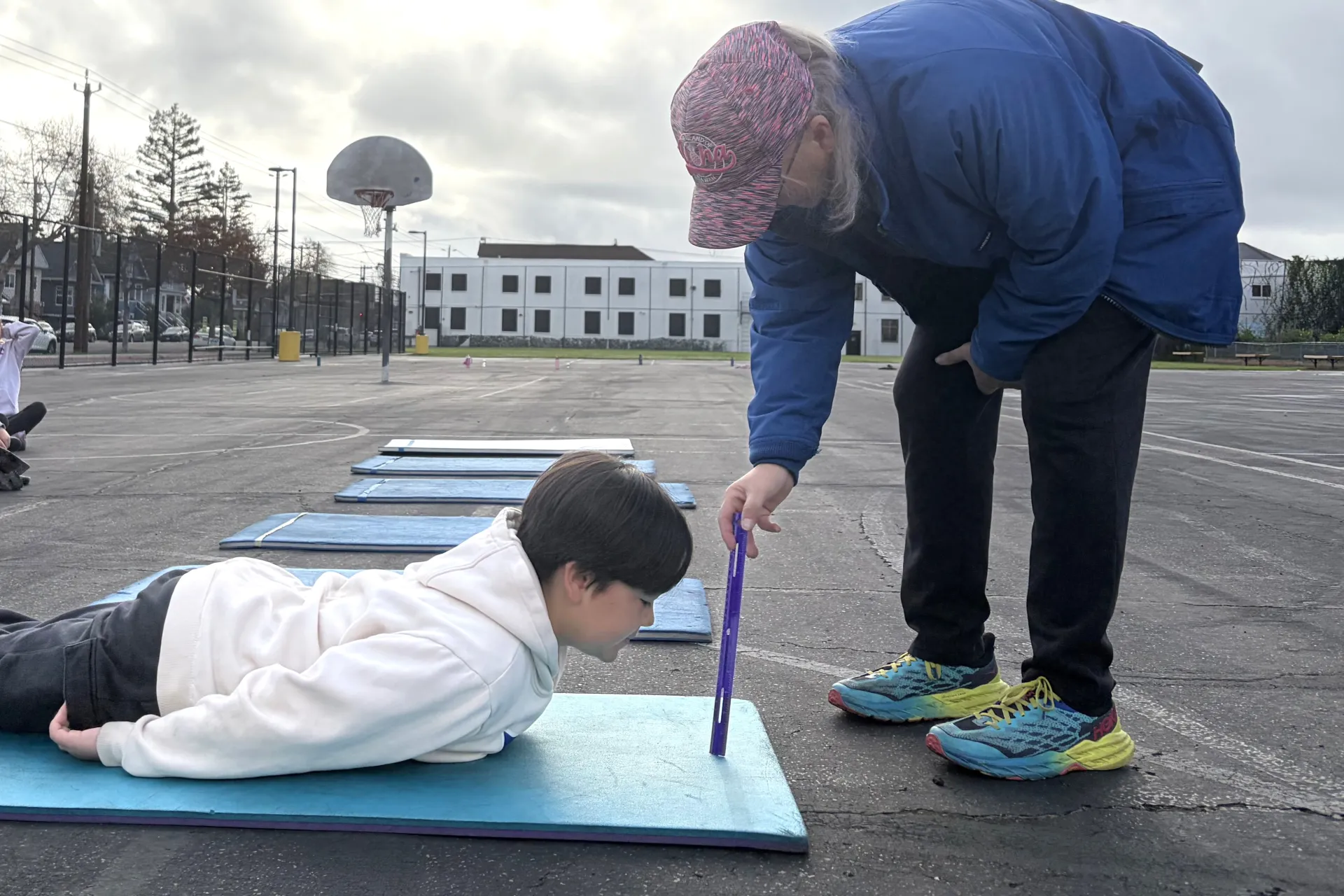 A physical education teacher stoops down with a ruler to measure a fifth grader's performance on a trunk lift at a school yard