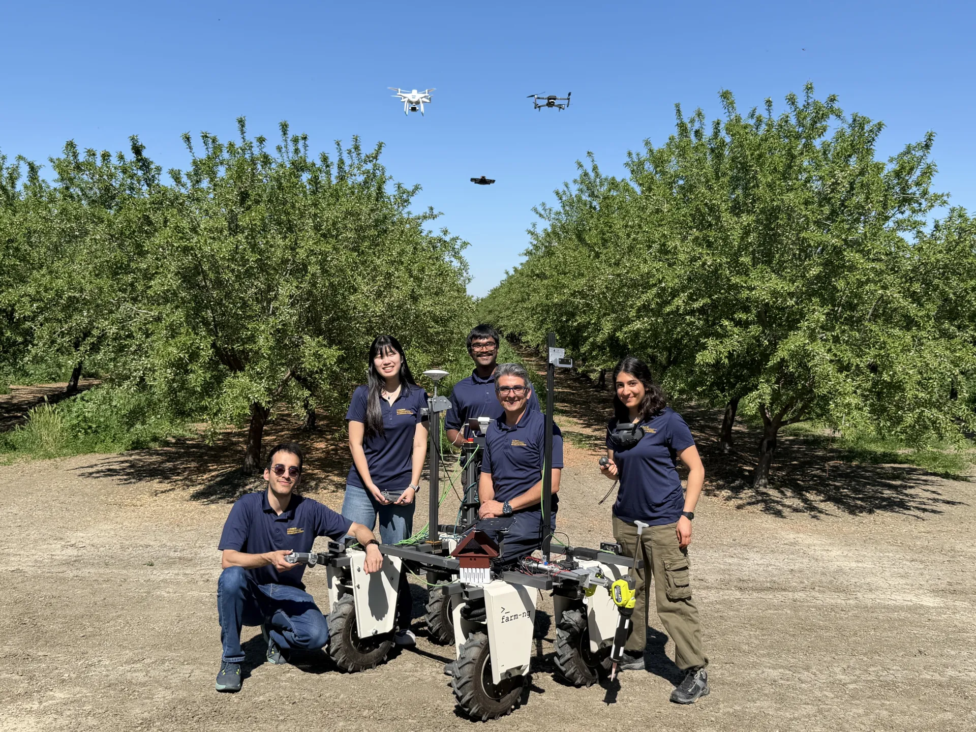A team of five from UC Davis, wearing matching shirts, pose by their ag robot in an orchard as three drones fly above