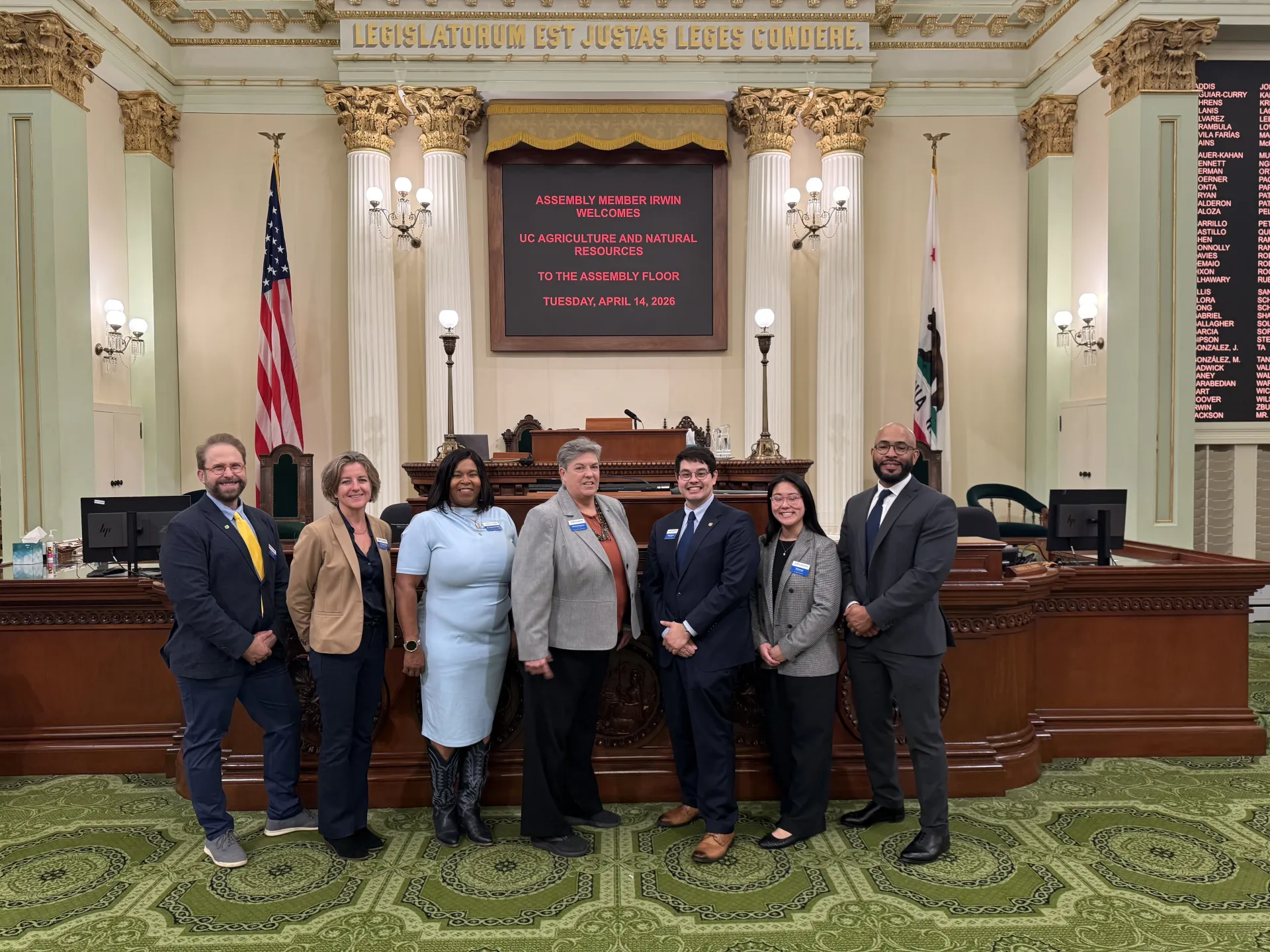 Seven people stand in a line in ornate chamber with pillars. Signs read: Legislatorum est justas leges condere. Assemblymember Irwin welcomes UC Agriculture and natural resources to the Assembly floor Tuesday, April 14, 2026