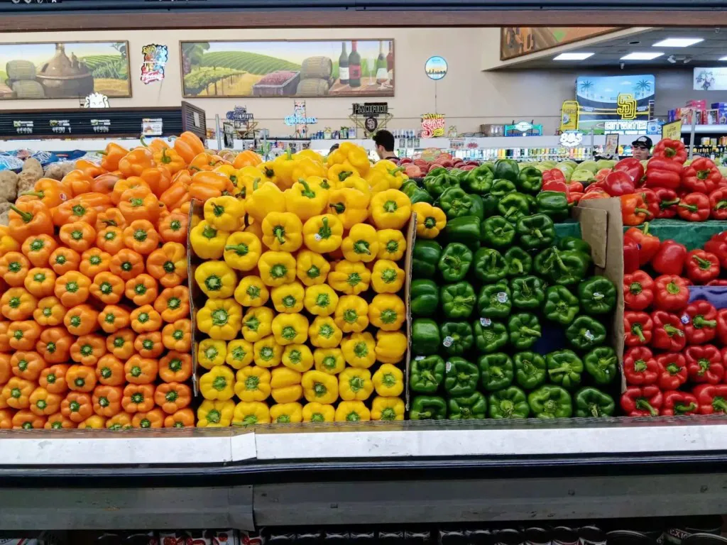 A supermarket produce display of neatly stacked bell peppers. From left to right, the peppers are organized in stacked columns by orange, yellow, green, and red. Store lights and decorative vineyard-themed signage is in the background.
