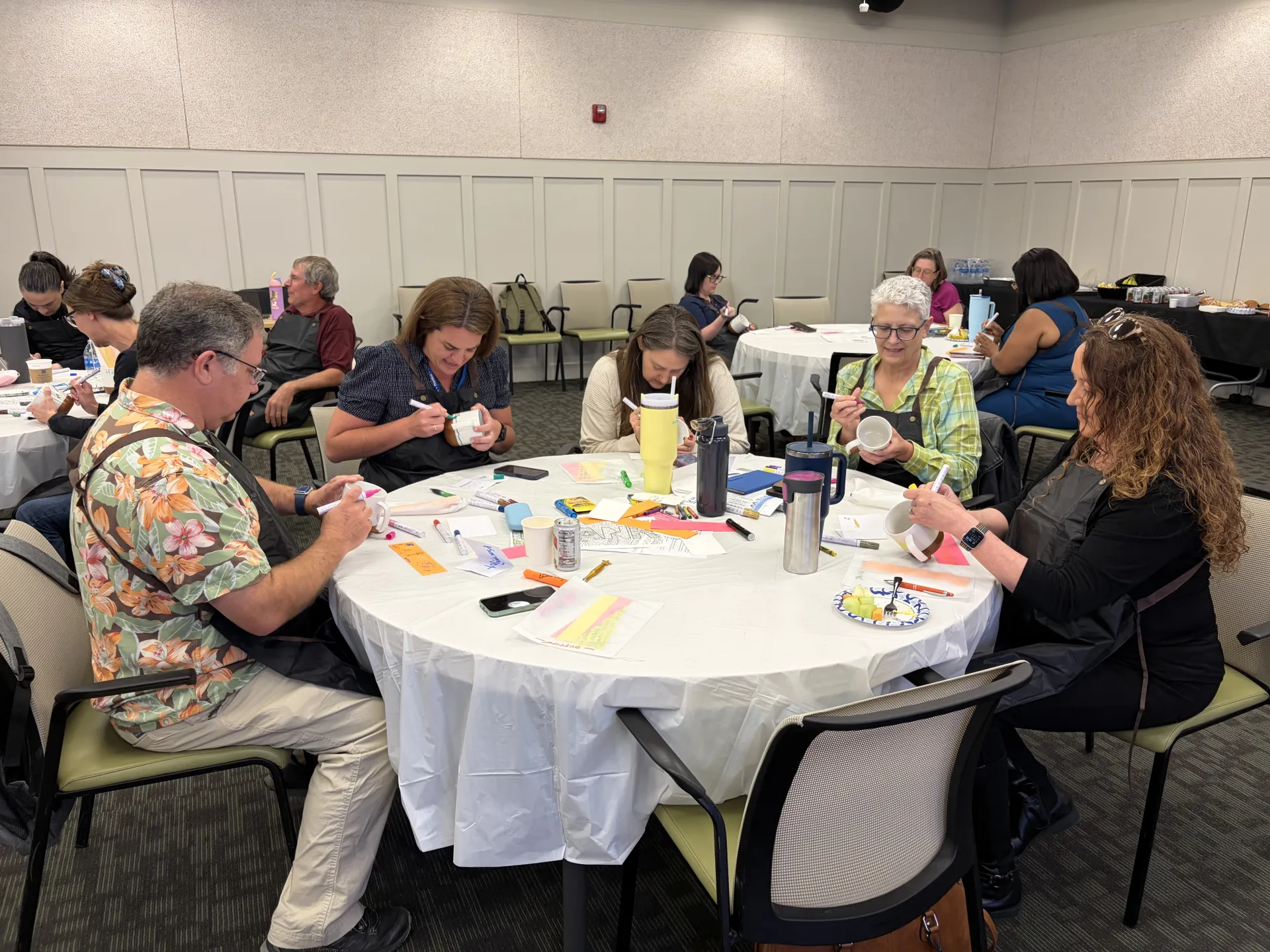 People sit at round tables drawing with colored pens on white coffee mugs
