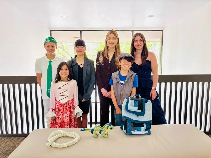 6 youth in their outfits they sewed for a fashion event on the couch in front of them are projects they made in their classes. A crocheted snake and upcycled backpack