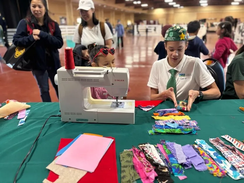 A 4-H youth volunteer sits behind a sewing machine, showing children how to work with colorful fabric scraps during a hands-on sewing activity. Families and visitors walk through the event space in the background.