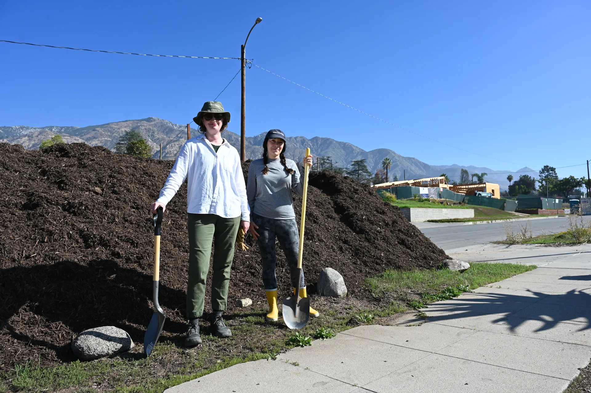 Two people with shovels stand in front of a pile of compost.