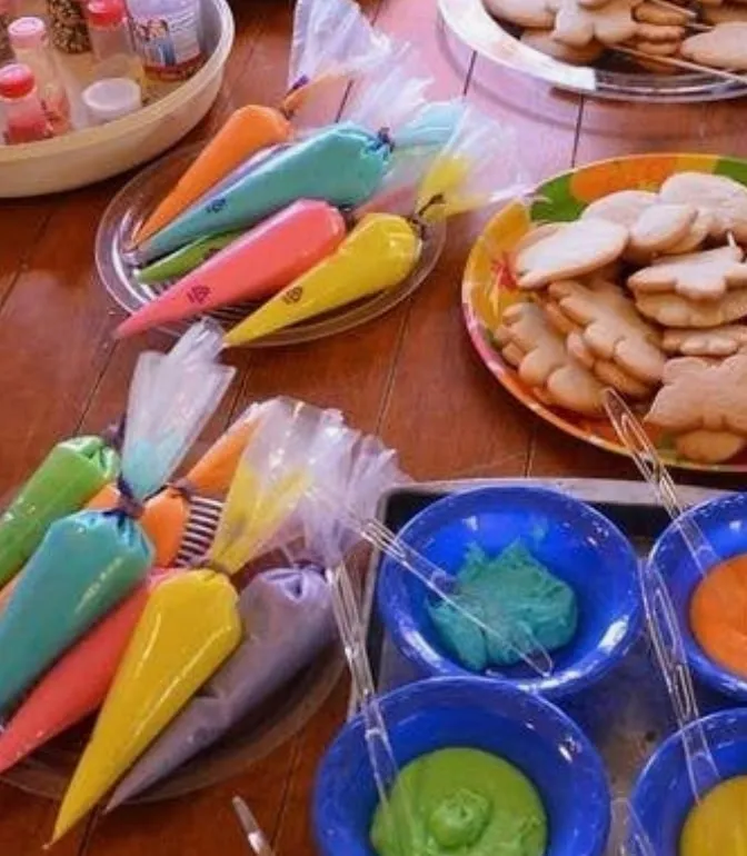 Colorful cookie decorating activity with plates of sugar cookies, bowls of brightly colored icing, and piping bags filled with pink, yellow, green, blue, orange, and purple icing.