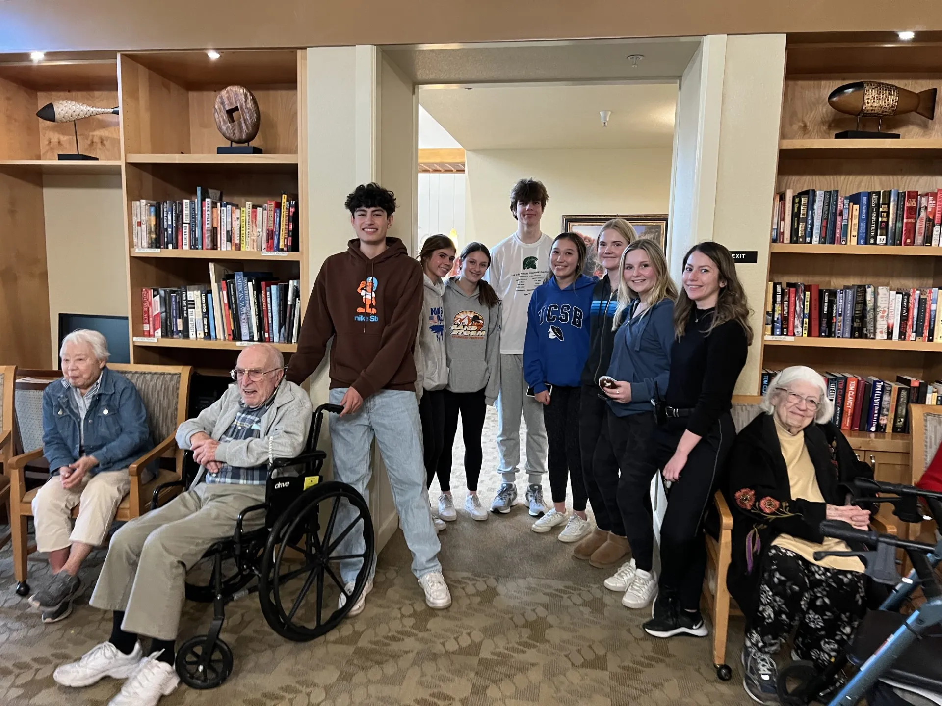 Group of teens and adults posing together in a cozy library or common room, standing and seated around two older adults in wheelchairs, with bookshelves filled with books in the background.