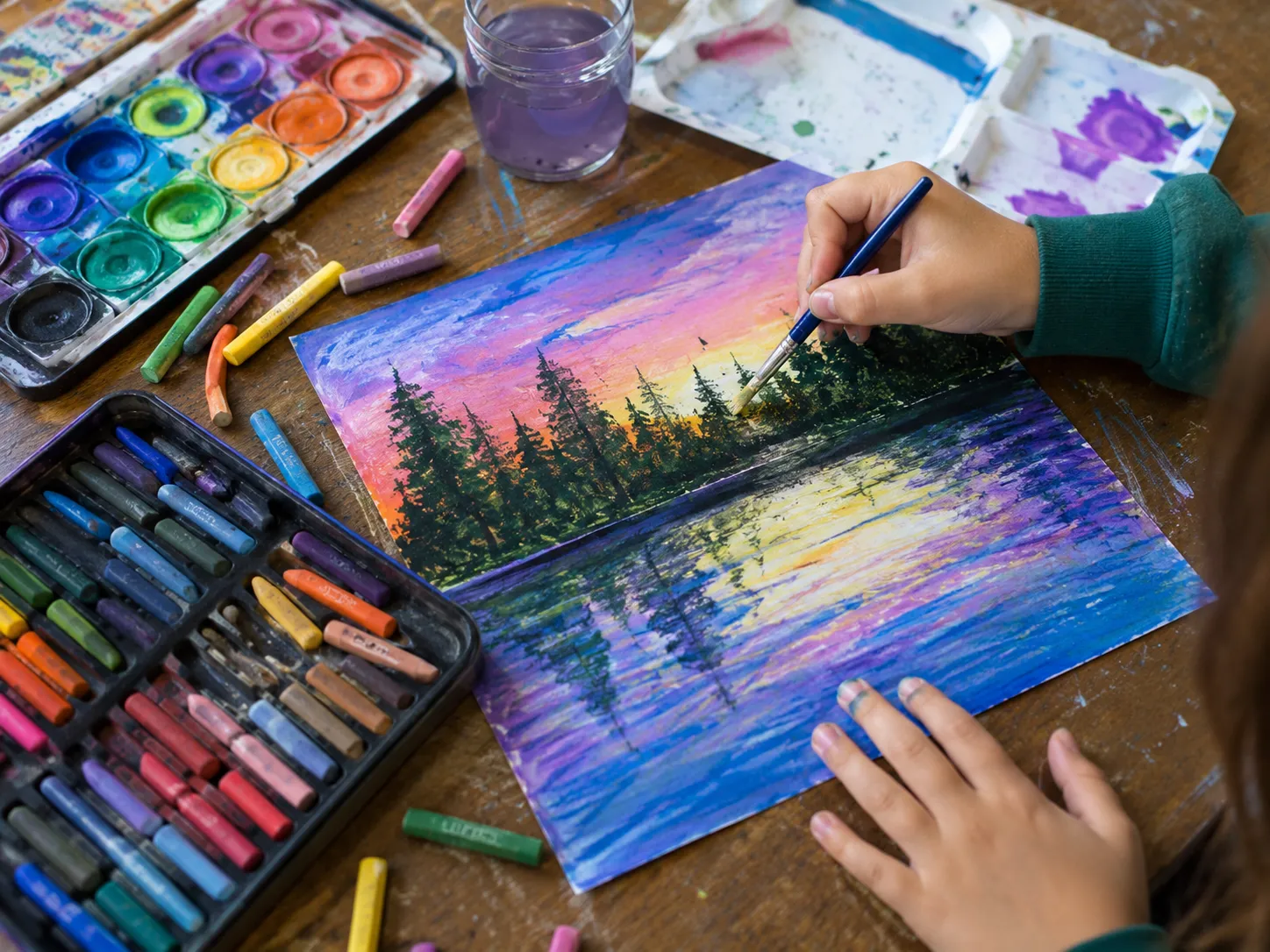 Close-up of a child painting a colorful sunset landscape while surrounded by watercolor paints, pastel sticks, brushes, and other art supplies on a wooden table.