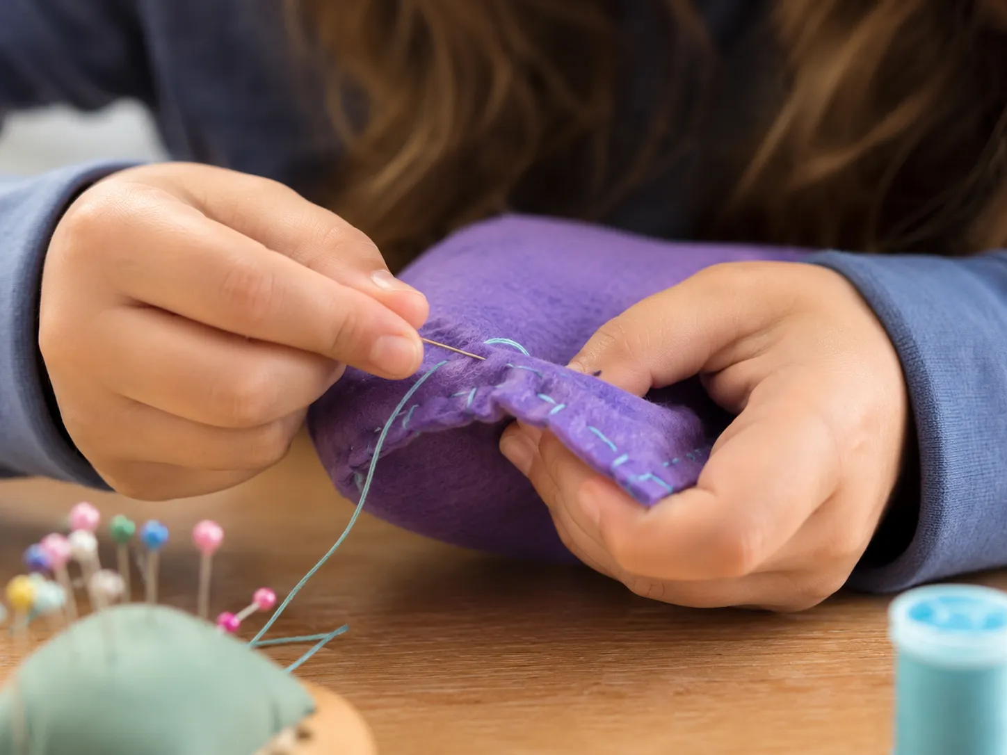 close-up of a girl hand sewing a purple piece of fabric a collection of needles and blue thread on the table.