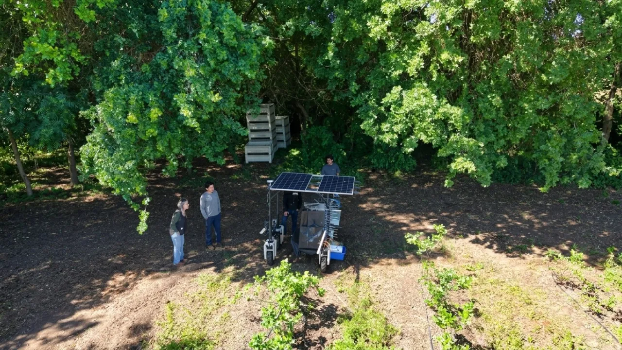 Four people look at an agricultural robot working at the edge of an orchard