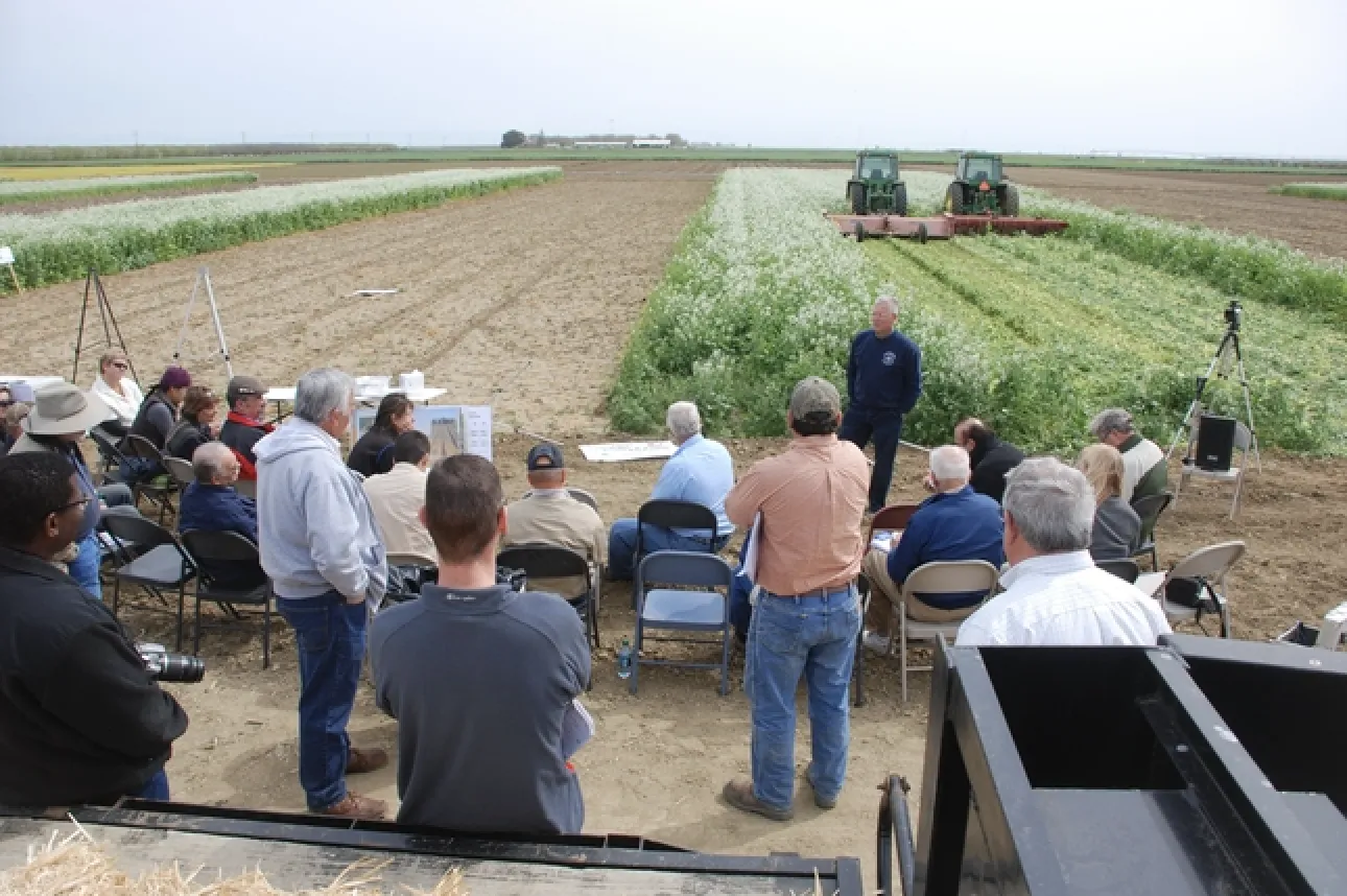 About 20 people sitting in folding chairs and standing on the edge of a crop field listen to a man speak. Two harvesters are running side by side in the field behind the speaker