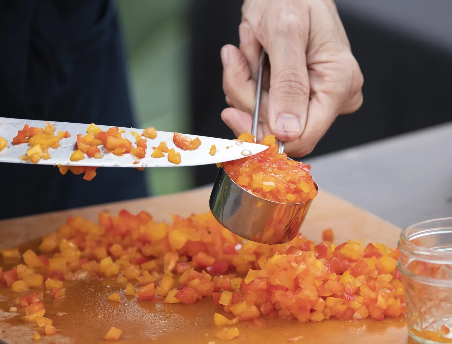 A person dicing orange peppers and using a knife to scoop the peppers into a measuring cup. 
