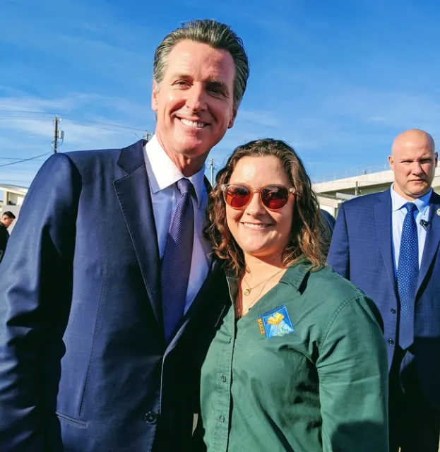 Gov. Newsom, wearing a dark suit, stands beside Katherine Uhde, who is wearing a green Master Gardener blouse.