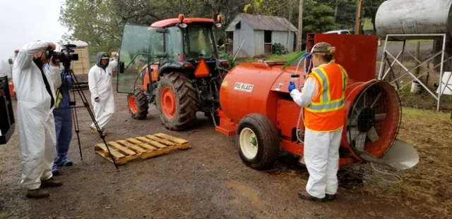 A photo showing a woman by an airblast sprayer being filmed while she measures flow rate from the nozzle.