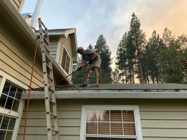 Ryan Tompkins stands on his roof raking moss, vegetative debris, and leaf litter from the gutters.