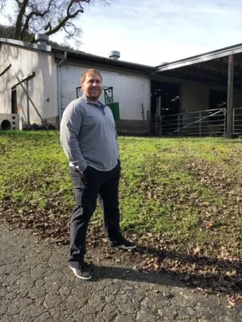 Keith Taylor stands outside on a muddy, grassy area with a building and tree behind him.
