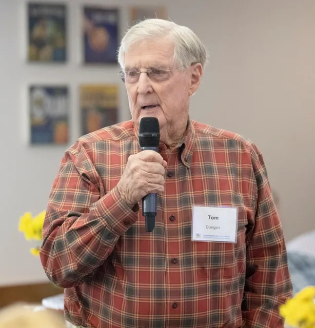 Tom Dungan, wearing a rust and brown plaid collared shirt, holds a microphone as he speaks.