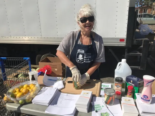 woman standing in front of a delivery truck and behind a table with lemons, papers, jarred vegetables, vinegar, salt and spices