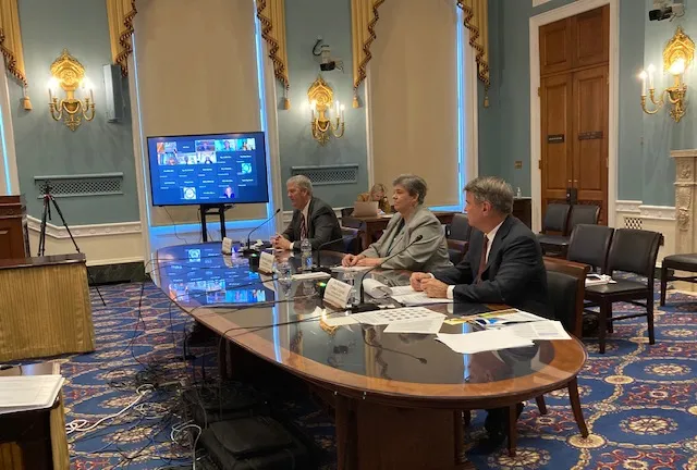 Glenda Humiston sits at a large oval table flanked by two men in dark suits. At one end of the table is a computer screen showing images of people joining the meeting via Zoom.