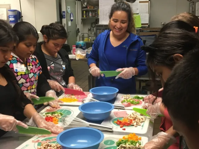 Youth standing at a counter with salad knives, chopping vegetables while a teacher in the center smiles at their work
