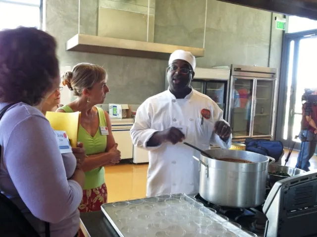 Two people listen as chef in white hat and uniform speaks while stirring large pot of soup.