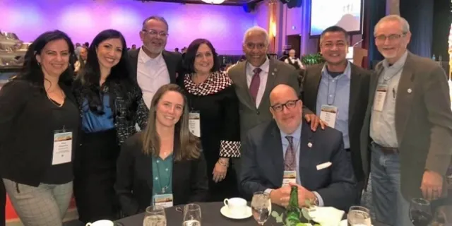 Nine people - 2 sitting and 7 standing - around a table in a conference hall.