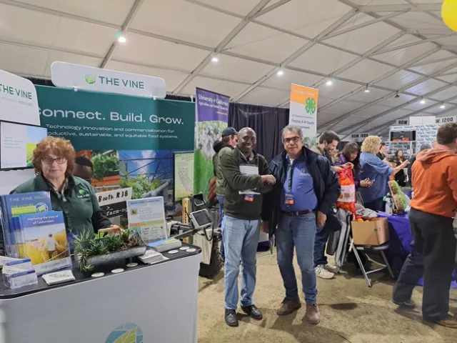 White stands behind a counter topped with handouts about The VINE. Behind Yaghmour, a man reaches into a bag labeled 