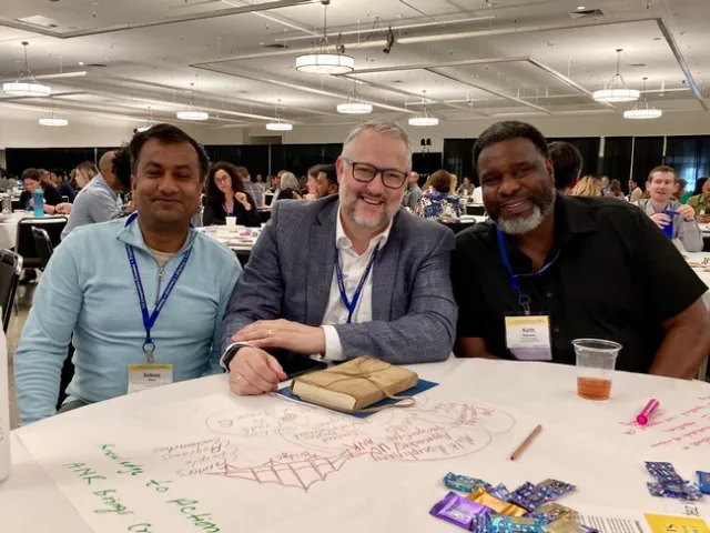 Three men sit at a round table covered with sketches and words written on white paper.