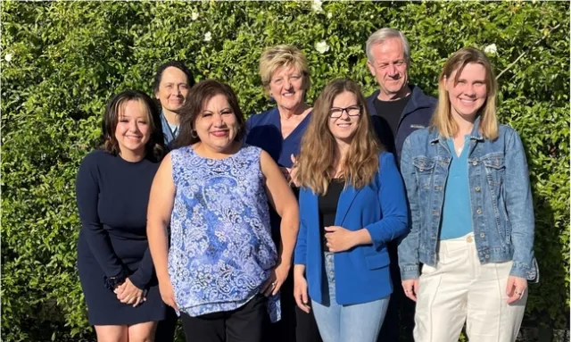Seven people pose standing in front of flowering hedge