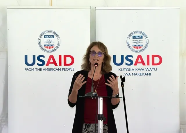 Mitcham speaks at a microphone in front of banners reading,