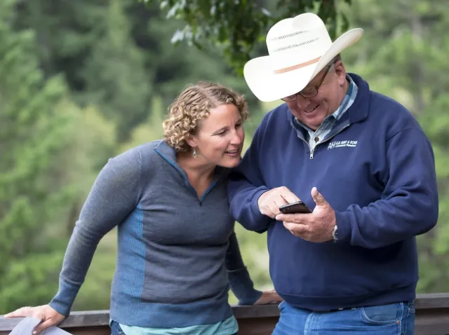 Lenya smiles and looks at whatever a man in a cowboy hat is pointing at on his phone.