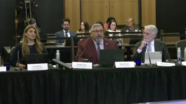 Three people sit at a long table facing the camera. A gallery of spectators sit behind them.