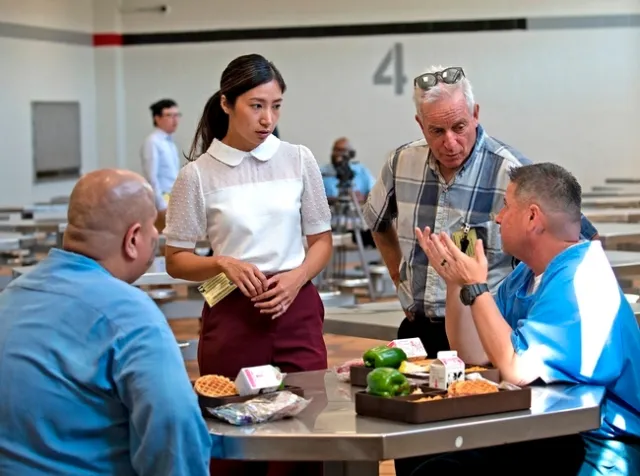 Heile Gantan of Impact Justice and Ron Strochlic of Nutrition Policy Institute (both standing) chat with residents wearing blue shirts sitting at an octagonal table about the quality of their food.