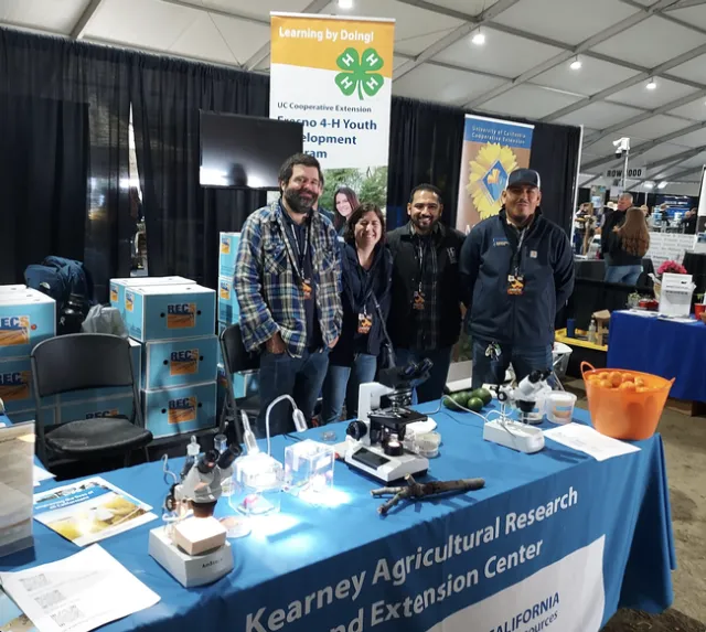 Four people stand behind a table covered with a blue tablecloth labeled Kearney Agricultural Research Extension Center and next to boxes labeled RECs