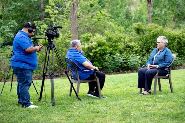 A man behind a camera records another man interviewing a woman while sitting down.
