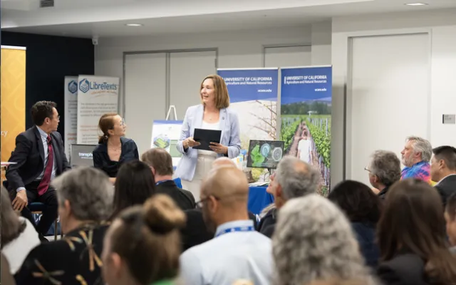 Leib and Park are sitting in front of a seated audience. Irwin is standing, holding a black notepad as she speaks to the group.