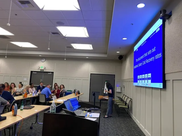 People sitting at conference room tables read the text on the PowerPoint screen: