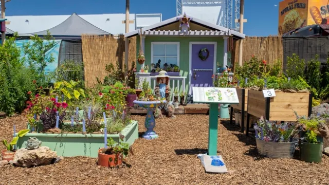 Plants in containers in front of a little house. A guide identifying the plant varieties sit atop a stand.