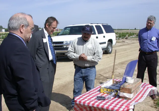 Manuel Jimenez gives Dynes samples of blueberries, which are displayed in plastic containers on a table with a red and white tablecloth on the edge of a field.
