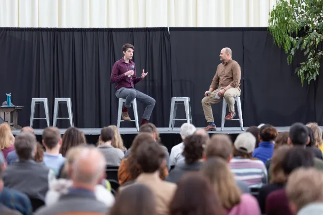 Swain sits on a stool on stage speaking to another man on stage