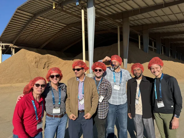 Seven people wearing hairnets pose in front of piles of almond hulls