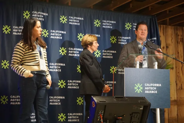Gabe Youtsey speaks at a podium while Karen Aceves and Karen Ross look on at AgTech Alliance press conference