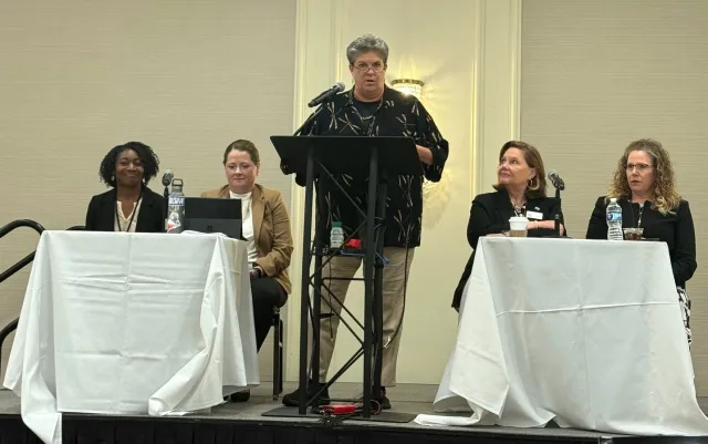 Glenda stands speaking between two tables of panelists