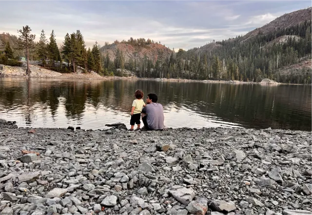 River rock in foreground, child and adult sit at the water's edge looking toward evergreens and hills