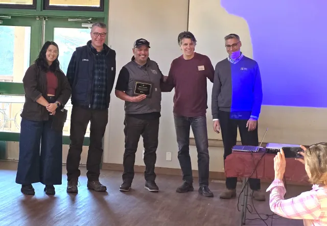 A woman and four men pose for a photographer as the man in center holds a plaque.