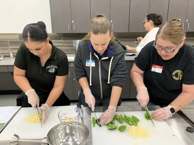 Three women cutting fresh garlic and green peppers on cutting boards in a kitchen