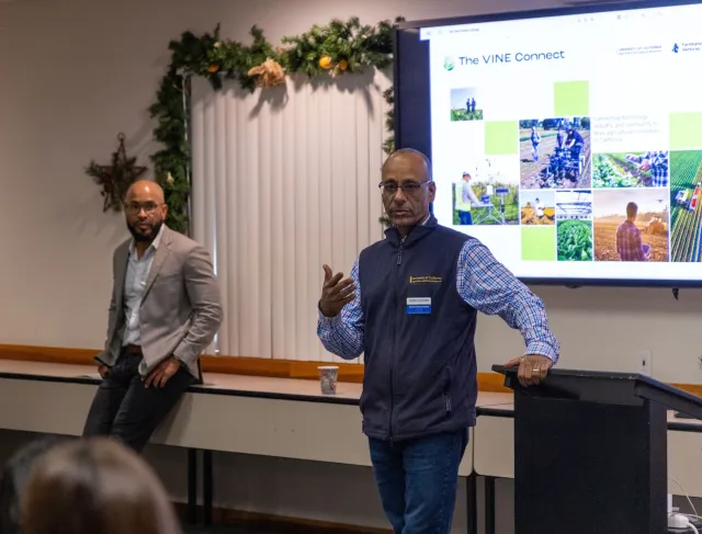 Two men stand in front of a PowerPoint slide that reads, "The VINE Connect" with images of people using technology in crop fields